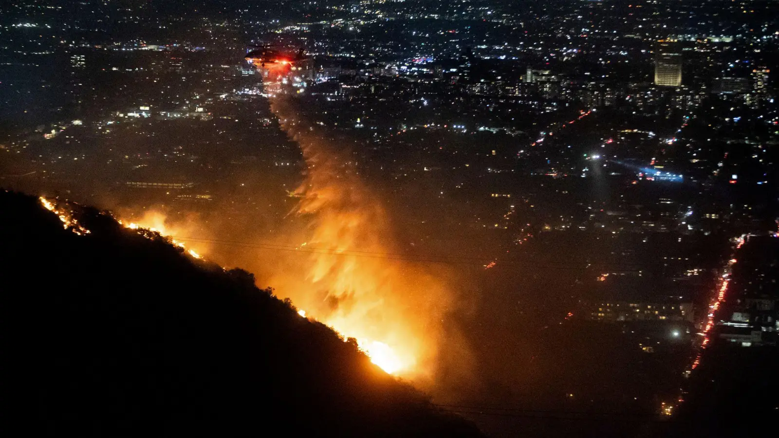 Ein Hubschrauber wirft Wasser auf das brennende Sunset Fire in den Hollywood Hills. (Bild: Ethan Swope/AP/dpa)