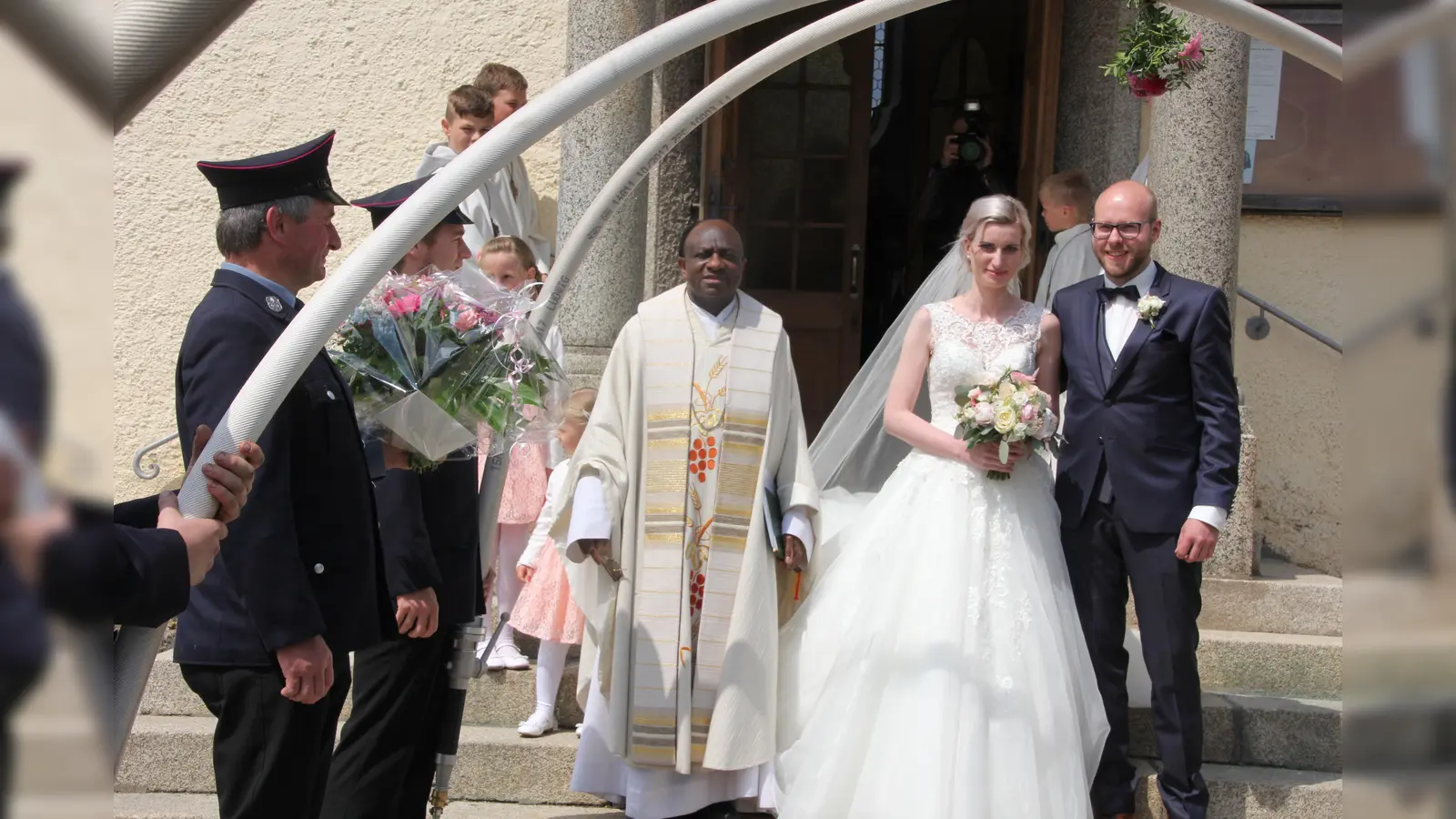 Luisa Schedl und Alex Wölfl wurden nach der Trauung in der Großkonreuther Kirche von der Feuerwehr Dippersreuth im Ehrenspalier erwartet. Links Pfarrer Dr. Charles Ifemeje. (Bild: kro)