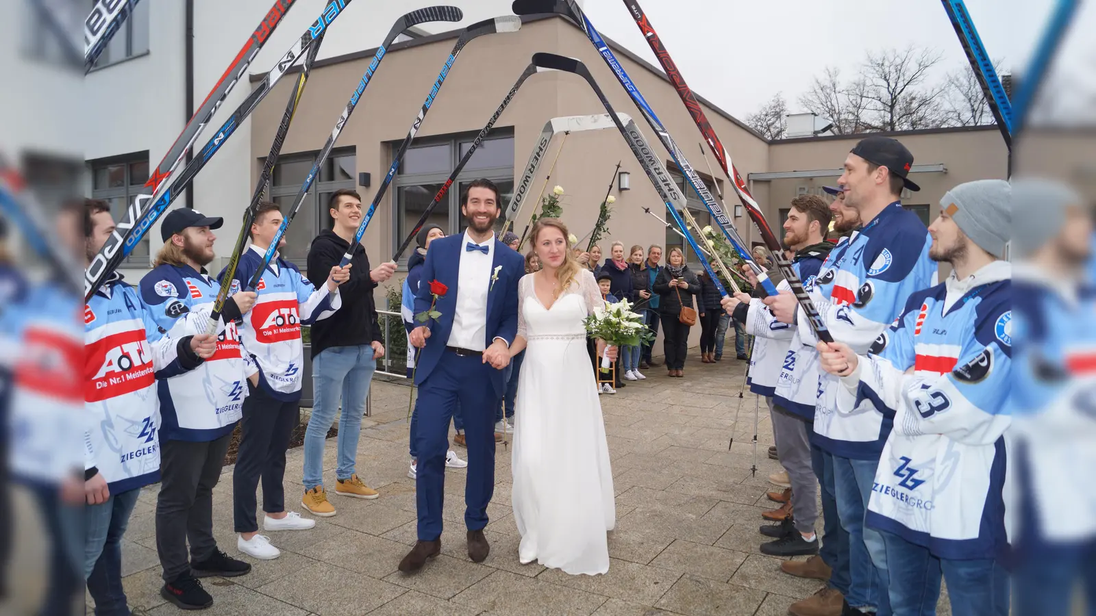 Christian Wolf und seine Frau Mareike schreiten nach ihrer standesamtlichen Hochzeit vor Bürgermeister Ernst Lenk im Schirmitzer Bürgersaal durch ein langes Gratulationsspalier von Freunden und Bekannten sowie den Eishockey-Spielern des EV Weiden. (Bild: du)