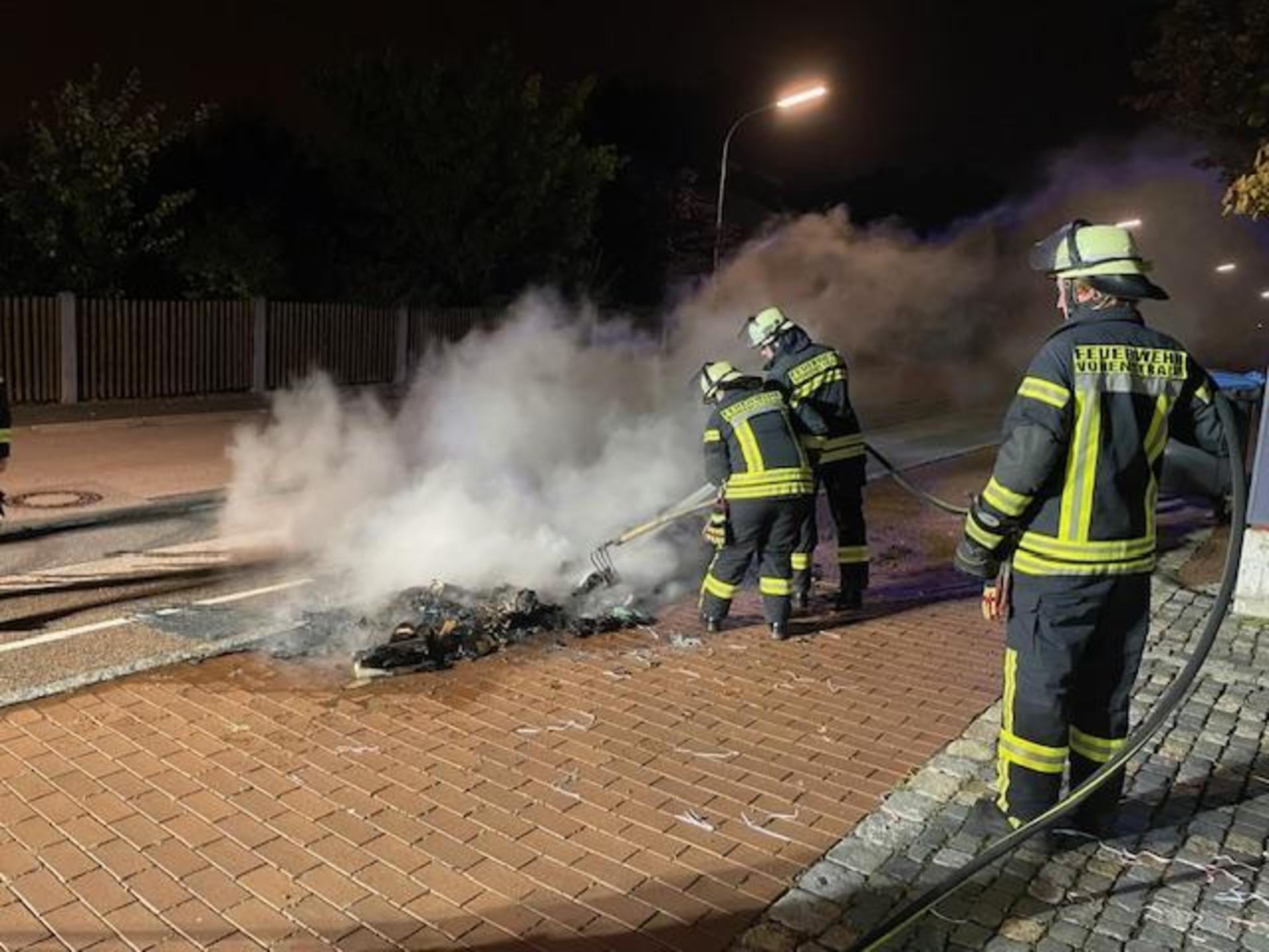 Die zur Abholung bereitgestellten Papiercontainer an der Pfalzgraf-Friedrich-Mittelschule brennen vollständig ab. Auch der Asphalt des Gehsteigs wird in diesem Bereich in Mitleidenschaft gezogen. Die Feuerwehrkräfte können nur mehr Nachlöscharbeiten durchführen.  (Bild: Anton Schwägerl/exb)