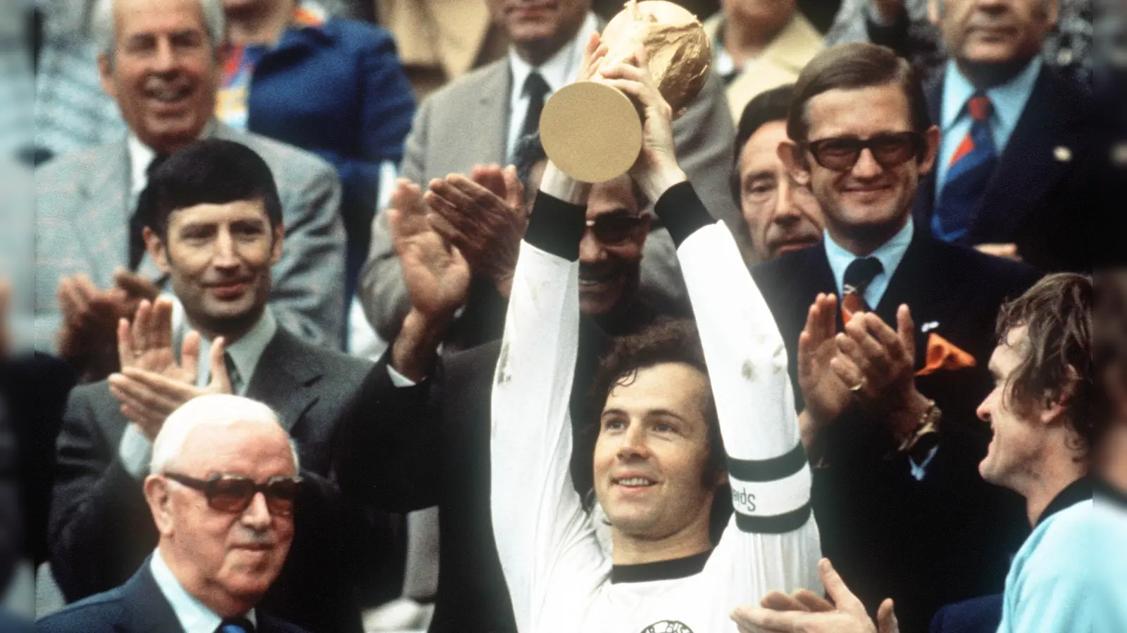 Franz Beckenbauer (M) hebt an der Seite von Torhüter Sepp Maier (r) und dem englischen FIFA-Präsidenten Stanley Rous (l) am 07.07.1974 im Münchner Olympiastadion nach dem Endspiel der Fußball-WM den eroberten WM-Pokal triumphierend hoch. Die deutsche Fußballnationalmannschaft gewann das Finale der Fußball-Weltmeisterschaft gegen die Niederlande mit 2:1. (Bild: Hartmut Reeh/dpa)