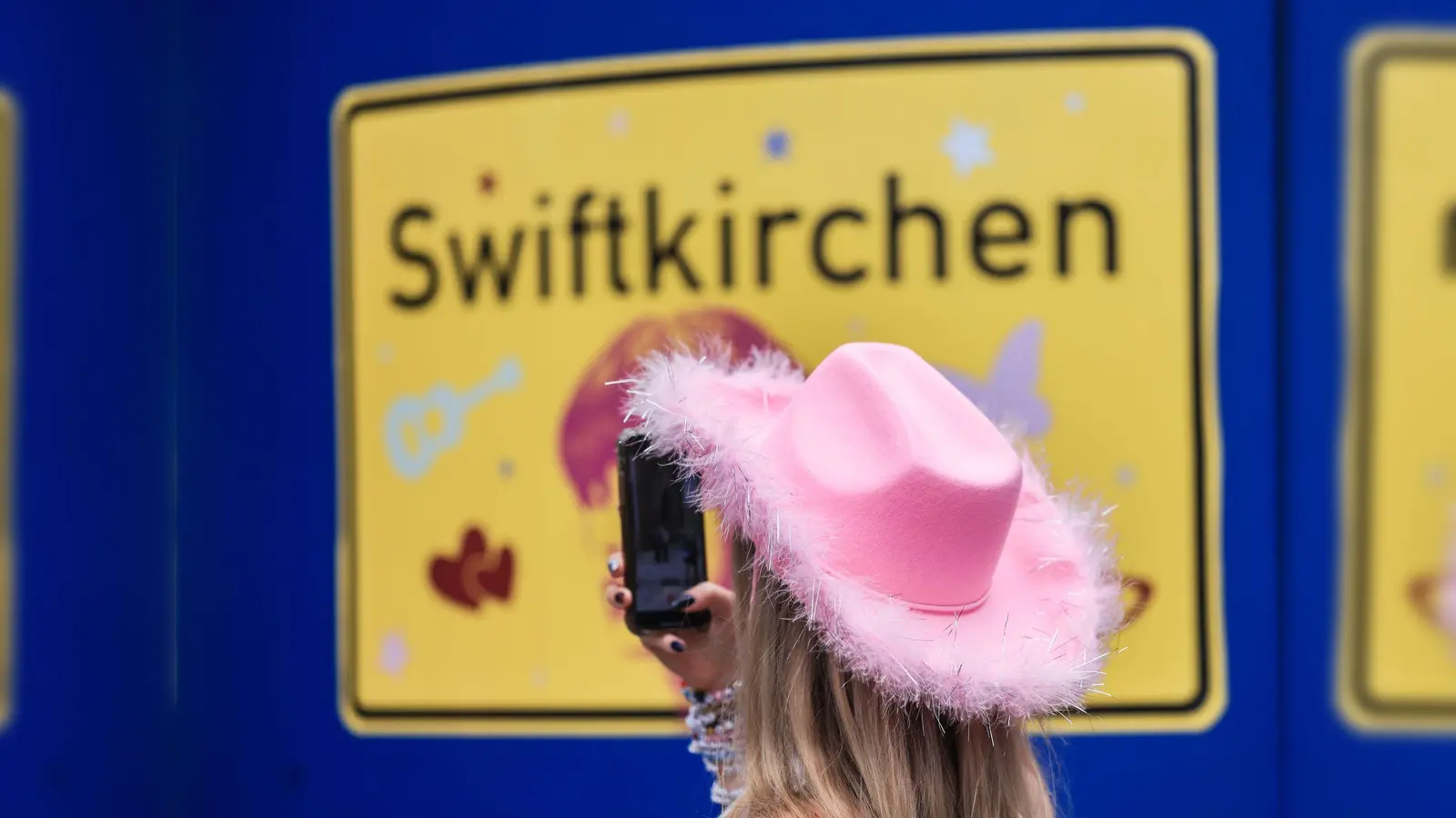 Ein Fan fotografiert vor dem Konzert der Sängerin Taylor Swift in Gelsenkirchen ein „Swiftkirchen“-Schild. Während des Konzerts am Abend zog der Datenverbrauch in den Handynetzen in der Veltins Arena kräftig an. (Bild: Oliver Berg/dpa)